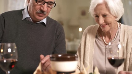 Tilting close-up shot of elderly Caucasian couple enjoying conversation and red wine over romantic dinner in hotel restaurant, toasting each other and chatting animatedly - Powered by Adobe