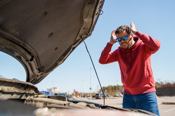 Photo of upset man with hands behind head next to open hood of broken car in daytime