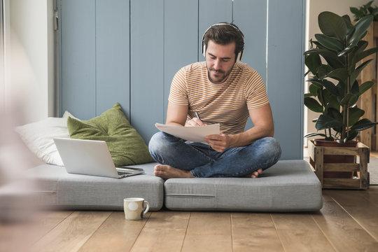 Young man sitting on mattress, taking notes, using headphones