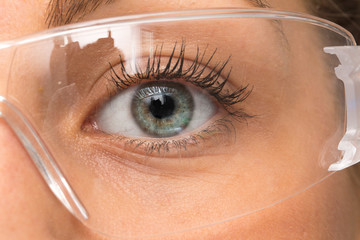 A closeup view on the greenish blue eye of a young laboratory technician wearing plastic safety glasses to protect from acid splashes and chemical burns, health and safety at work.