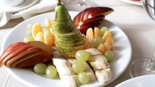 Close-up panning object shot of fruit platter sitting on table in restaurant, with sliced pear, apple, banana, grapes and tangerine, with empty wine glasses beside plate on beige tablecloth
