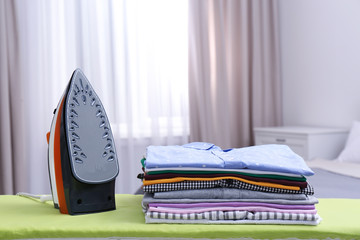 Modern electric iron and clean folded clothes on board in bedroom. Laundry day