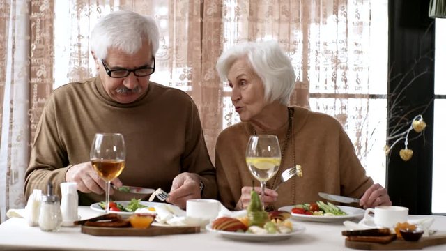 Medium shot of senior Caucasian husband and wife, wearing beige jumpers, sitting together at table by window, eating healthy breakfast and discussing something, looking disappointed and dismayed