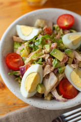 Traditional american cobb salad with chopped avocado, tomatoes, hard boiled egg, roasted chicken and greens for a healthy lunch, served on wooden table with soft light