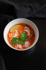 Top view of oriental tomato noodle soup with champignon mushrooms, rice noodle, parsley and redish, served on total black background