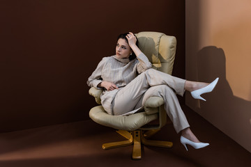 trendy model sitting on armchair, crossing legs, looking away