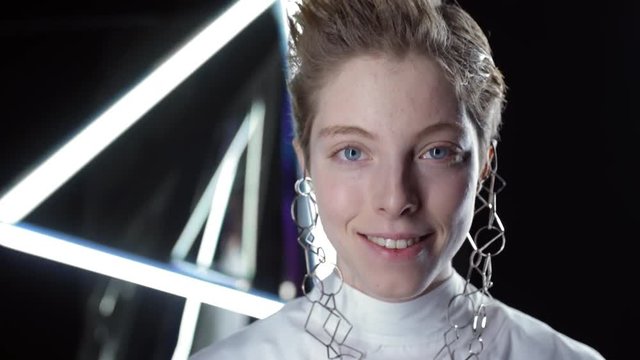 Close up of teenage Caucasian girl with short hair and no makeup wearing trendy futuristic earrings smiling and looking at camera against black background with neon lamps