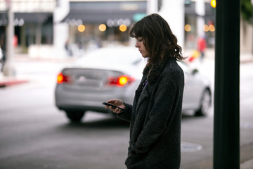 Female pedestrian waiting for a rideshare.  She is sharing her gps location via cellphone app so the driver can pick her up in the city.  Cars are blurred to obscure make model and license plates.