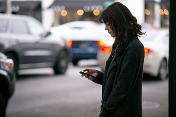 Female pedestrian waiting for a rideshare.  She is sharing her gps location via cellphone app so the driver can pick her up in the city.  Cars are blurred to obscure make model and license plates.