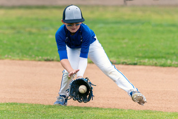 Youth baseball player in blue uniform fielding a ground ball into his glove in the infield during a...