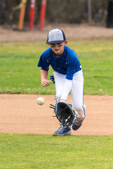 Obraz premium Youth baseball player in blue uniform keeping eyes on a ground ball into his glove in the infield during a game.
