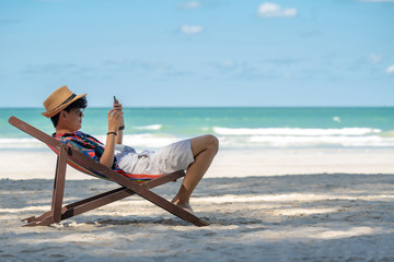Young asian adult men traveler in casual wear relaxing and lying down on beach chair on tropical island beach in summer holidays vacation travel trip using smartphone for text, selfie or take a photo