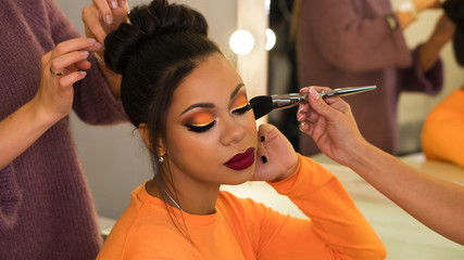 Hairstylist and make-up artist makeup and updo for stylish young African-American woman in beauty parlor. Rich colors. Party image. Beauty industry - obrazy, fototapety, plakaty