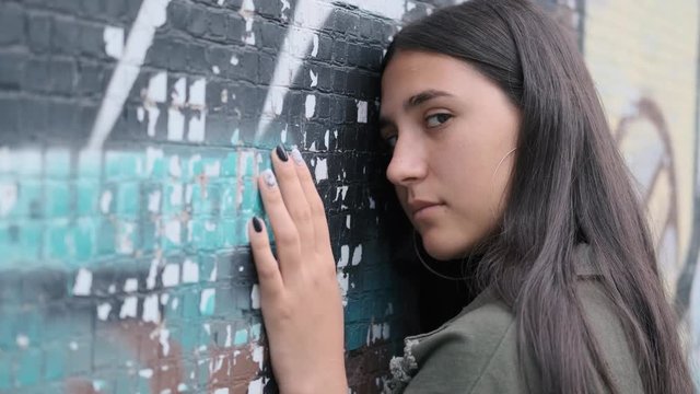 sad young brunette girl stands with her hand down and leaning her head on the wall with graffiti turns to look at the camera and runs her hand down the wall