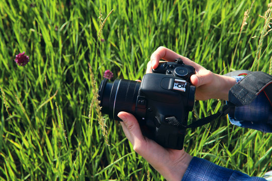 The hands of the girl hold the camera on the background of green grass. Cropped shot, horizontal, free space on the left. The concept of a hobby and profession.