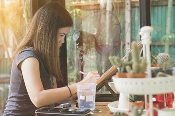 Beautiful woman embodying her ideas on paper with help of paintbrush and watercolors, interior of coffee cafe on natural background.