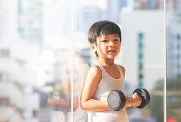 Little boy is working out with dumbbell by the windows city.