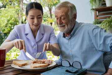 senior man happiness with smiling nurse, takes care breakfast at balcony near garden at nursing home