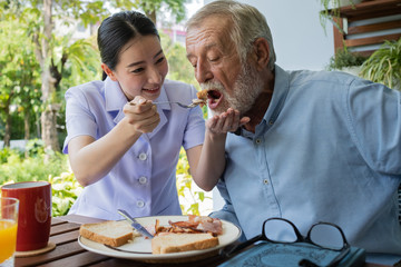 senior man happiness with smiling nurse, takes care breakfast at balcony near garden at nursing home