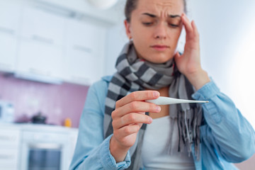Upset sick woman having high fever and suffering from strong headache during flu. Holding a medical thermometer