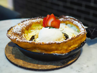 Delicious and beautiful Dutch baby pancake with vanilla ice cream topped with fresh strawberry, crumble, chocolate sauce and icing powder on preheat Cast-iron skillet. Selective focus.