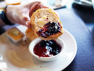 Woman hands spread blueberry jam over butter with a wooden spatula on side of baked scone. selective focus.