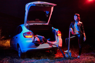 Female bandit near car with a shovel and a dead or drunk guy in the trunk at night. Photoshoot...