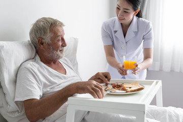 senior man with smiling nurse, takes care breakfast and discussion and cheer on bed at nursing home