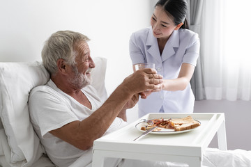 senior man with smiling nurse, takes care breakfast and discussion and cheer on bed at nursing home