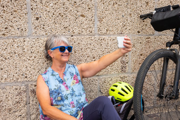 Happy senior woman with gray hair takes a selfie sitting against a wall. Resting after a bicycle excursion. Healthy lifestyle for a retired people