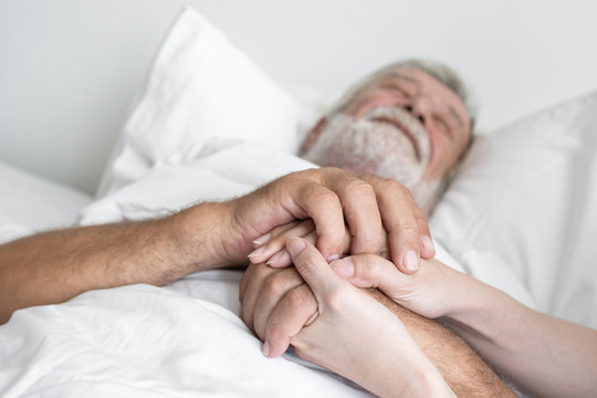 hand of senior man with smiling nurse, takes care and cheer on bed at nursing home
