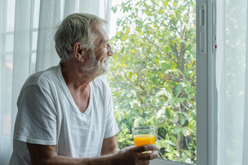 Fototapeta na wymiar senior man hold glass of orange juice and thinking and looking outside of window in white room