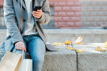 Autumn shopping. Cropped shot of man sitting on wall with packages, using smartphone. Fall urban...