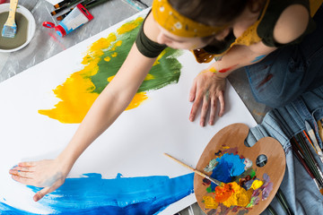 Finger painting hobby. Top view of female artist sitting on floor, using blue and yellow colors to...