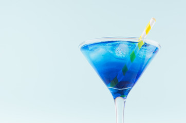 Summer cold blue lagoon drink with ice cube, yellow striped straw in elegant martini glass on pastel mint background, closeup, top part.