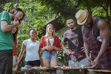 Group of happy friends standing eating and drinking beers at barbecue dinner camping in nature and having meal together outdoor as summer lifestyle, food and friendship concept