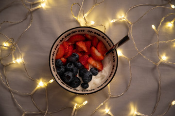 Top side view of a bowl with yogurt, strawberries and blueberries over grey sheets with lights