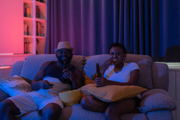 Naklejka premium Smiling man and woman holding beer excited with emotional in the dark night neon color light party environment background at home