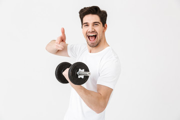 Emotional happy excited young man posing isolated over white wall holding dumbbell make exercise pointing.