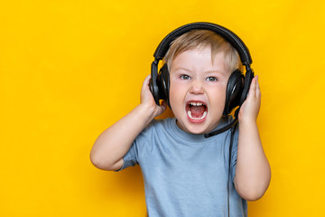 Little boy in modern headphones shouting, standing on yellow background