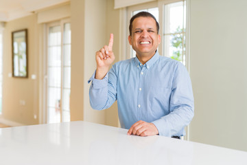 Middle age man sitting at home showing and pointing up with finger number one while smiling confident and happy.