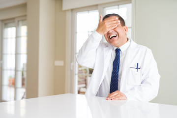 Fototapeta premium Middle age doctor man wearing medical coat at the clinic smiling and laughing with hand on face covering eyes for surprise. Blind concept.