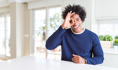Young african american man wearing casual sweater sitting at home doing ok gesture with hand smiling, eye looking through fingers with happy face.