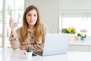Beautiful young woman working with computer takes a break to drink glass of water with a confident expression on smart face thinking serious