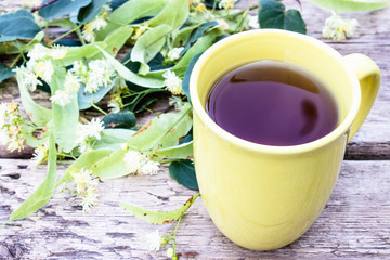 Linden tea in a yellow cup stands near linden flowers against the background of old boards.