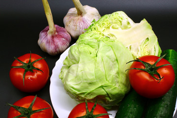 On a black background - two garlic, four tomatoes, one cabbage, two green cucumbers. Not all vegetables fit in the plate