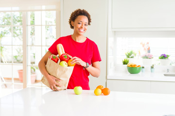 Young beautiful african american woman holding paper bag full of fresh healthy groceries leaving vegetables on the table