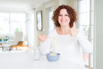 Senior woman eating asian noodles using chopsticks happy with big smile doing ok sign, thumb up with fingers, excellent sign