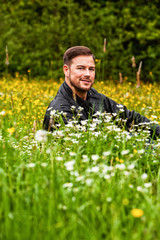 Junger Mann mit Lederjacke, sitzend auf einer Blumenwiese mit Blick in die Kamera