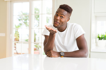 Handsome african american man on white table at home looking at the camera blowing a kiss with hand on air being lovely and sexy. Love expression.